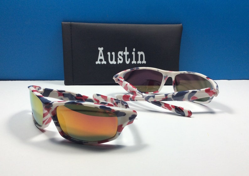 May include: A pair of white sunglasses with a red, white and blue camouflage pattern. The sunglasses have mirrored lenses and are folded in half. The sunglasses are sitting on a white surface next to a black case with the word "Austin" printed on it.