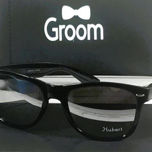 May include: Black sunglasses with a black frame and dark lenses. The sunglasses have the word "Hubert" printed on the right lens.