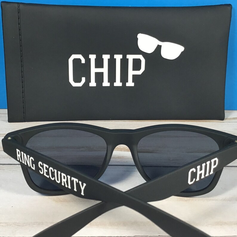 Op de afbeelding: Matzwarte zonnebril met donkere glazen, voorzien van de witte tekst RING SECURITY op &eacute;&eacute;n pootje en CHIP op het andere. Achter de zonnebril staat een bijpassend zwart rechthoekig etui met de witte tekst CHIP en een afbeelding van een zonnebril.