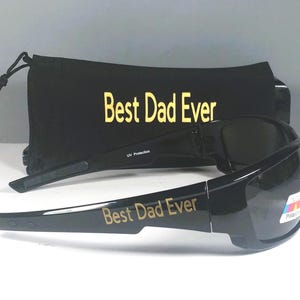 Puede incluir: Gafas de sol negras con lentes oscuras y el texto Best Dad Ever impreso en dorado en la patilla. Detr&aacute;s de las gafas hay una bolsa negra con cord&oacute;n a juego, que tambi&eacute;n presenta el texto Best Dad Ever en letras doradas.