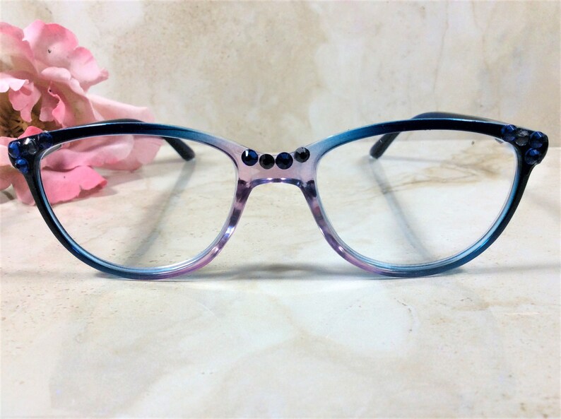 Puede incluir: Gafas de estilo ojo de gato con montura degradada azul y morada. La montura est&aacute; adornada con pedrer&iacute;a azul oscuro en las esquinas y el puente. Las gafas tienen lentes transparentes y varillas negras.