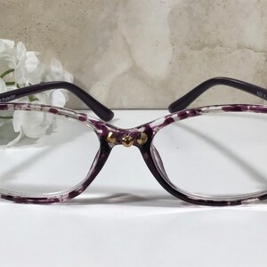 Puede incluir: Gafas con montura transparente con un estampado de lunares morados y patillas moradas oscuras. Las monturas est&aacute;n adornadas con adornos dorados. Las gafas tienen forma de ojo de gato y est&aacute;n dise&ntilde;adas para la correcci&oacute;n de la visi&oacute;n.