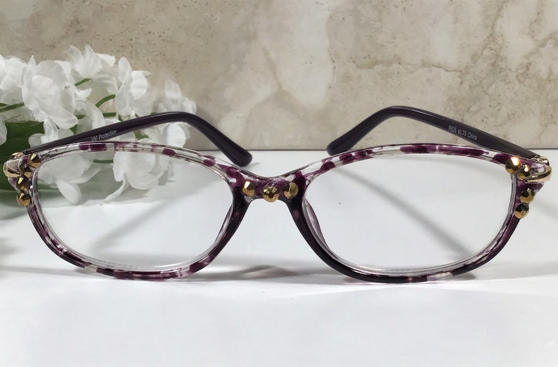 Puede incluir: Gafas con montura transparente con un estampado de lunares morados y patillas moradas oscuras. Las monturas est&aacute;n adornadas con adornos dorados. Las gafas tienen forma de ojo de gato y est&aacute;n dise&ntilde;adas para la correcci&oacute;n de la visi&oacute;n.