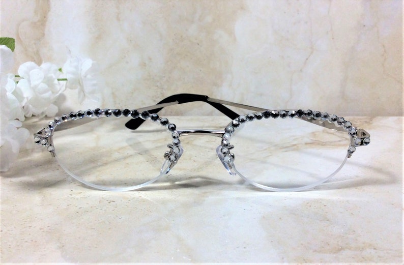 Pu&ograve; includere: Occhiali con montatura argentata e lenti trasparenti, adornati con strass scintillanti lungo il bordo superiore e sulle aste. Gli occhiali hanno un design delicato ed elegante, perfetto per aggiungere un tocco di glamour. La montatura &egrave; argentata con aste nere.
