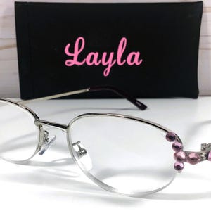 May include: Silver-framed eyeglasses with clear lenses and pink gemstone accents on the temples. A black case with the name "Layla" in pink script is behind the glasses. The glasses have a modern, elegant design.