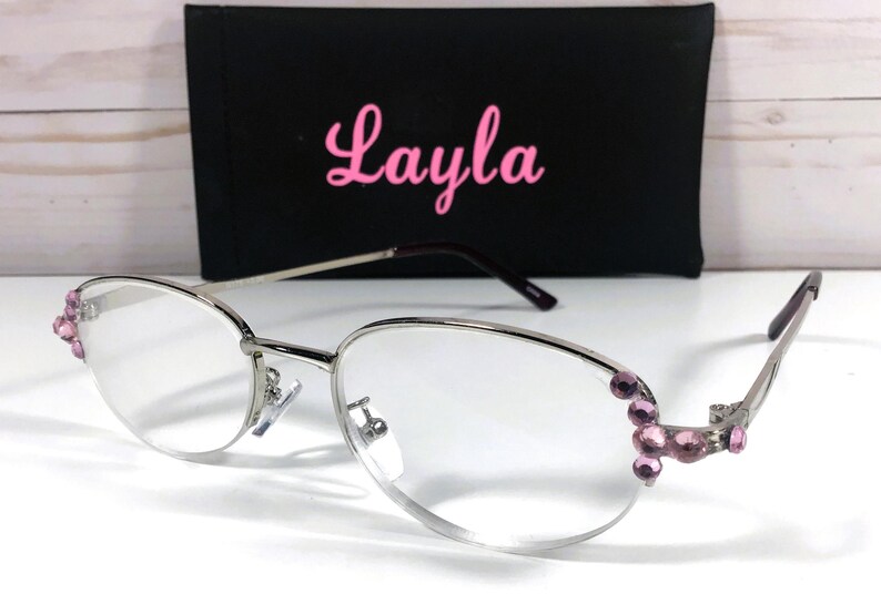 May include: Silver-framed eyeglasses with clear lenses and pink gemstone accents on the temples. A black case with the name "Layla" in pink script is behind the glasses. The glasses have a modern, elegant design.