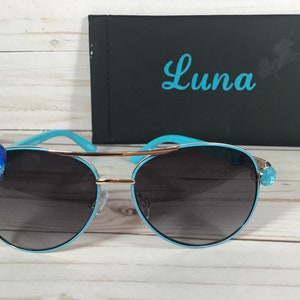 Pu&ograve; includere: Occhiali da sole aviator blu con dettagli dorati e decorazioni a forma di cuore blu sulle aste. Gli occhiali da sole sono in una custodia con la scritta "Luna" stampata su di essa.
