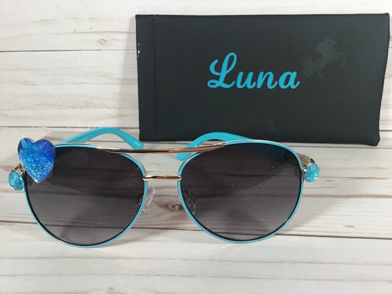 Pu&ograve; includere: Occhiali da sole aviator blu con dettagli dorati e decorazioni a forma di cuore blu sulle aste. Gli occhiali da sole sono in una custodia con la scritta "Luna" stampata su di essa.