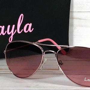 May include: Rose gold aviator sunglasses with gradient pink-tinted lenses. The right lens features the white text Layla. Behind the sunglasses is a black pouch with the name Layla printed in large pink script lettering.