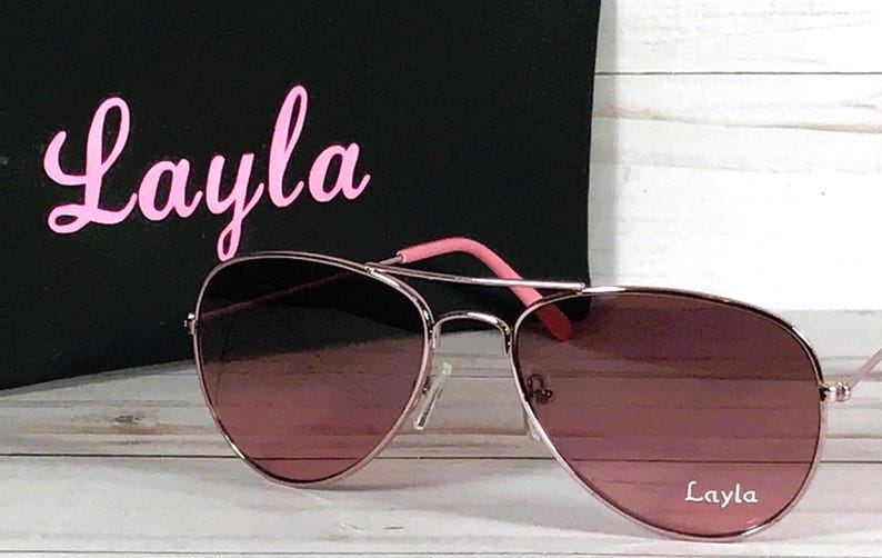 May include: Rose gold aviator sunglasses with gradient pink-tinted lenses. The right lens features the white text Layla. Behind the sunglasses is a black pouch with the name Layla printed in large pink script lettering.