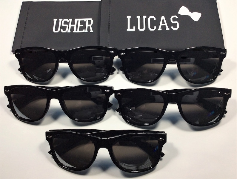 May include: Four pairs of black sunglasses with dark lenses. The sunglasses are arranged in two rows of two. The top row has the words "USHER" and "LUCAS" on the boxes behind them.