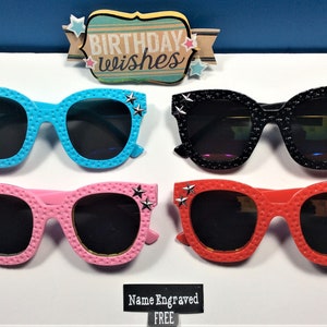 Puede incluir: Cuatro pares de gafas de sol con detalles de estrellas. Las gafas de sol son de color azul, negro, rosa y rojo. El texto "Birthday Wishes" es visible sobre las gafas de sol.