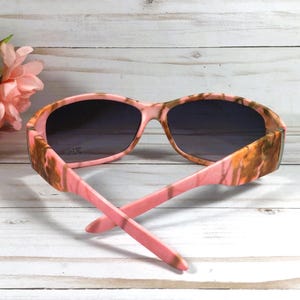 May include: Pink and brown camouflage sunglasses with dark lenses. The sunglasses are on a white wooden surface.