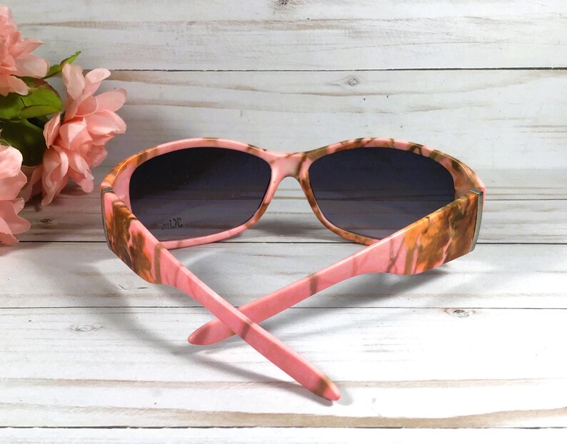 May include: Pink and brown camouflage sunglasses with dark lenses. The sunglasses are on a white wooden surface.