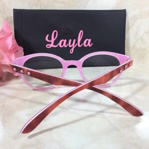 May include: Pink eyeglasses with brown arms and decorative studs. The glasses are paired with a black case with the name "Layla" in pink script. Pink flowers are in the background.
