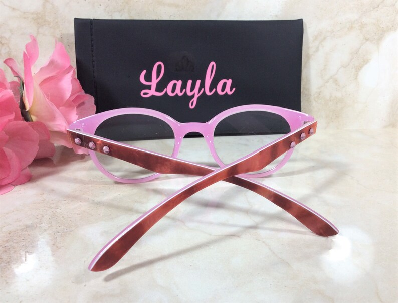 May include: Pink eyeglasses with brown arms and decorative studs. The glasses are paired with a black case with the name "Layla" in pink script. Pink flowers are in the background.