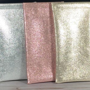 May include: Three glittery eyeglass cases in silver, rose pink, and gold. The cases are rectangular with a soft, textured appearance. The cases are standing upright, side by side.