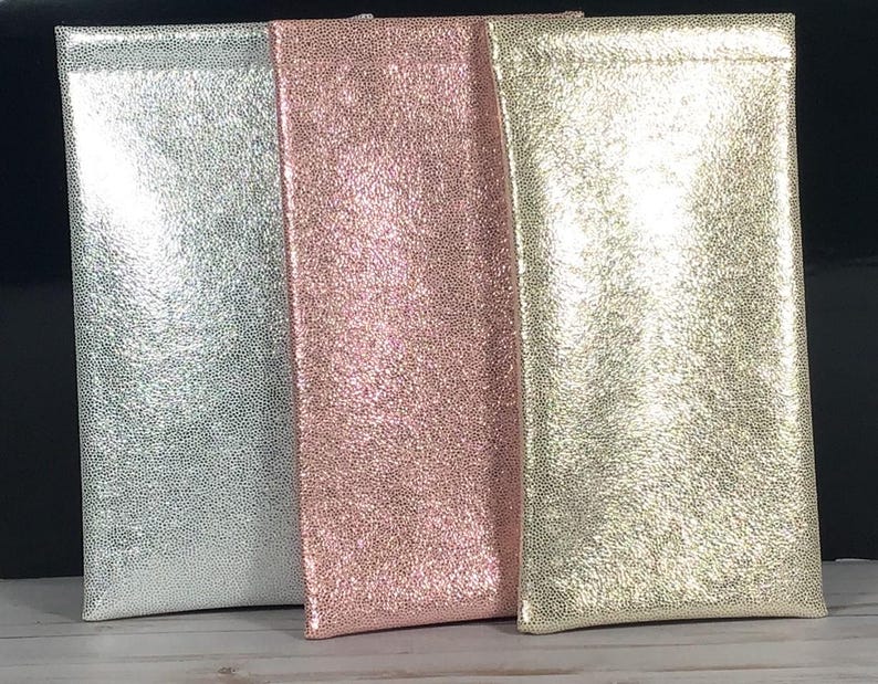 May include: Three glittery eyeglass cases in silver, rose pink, and gold. The cases are rectangular with a soft, textured appearance. The cases are standing upright, side by side.