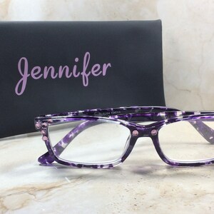 K&ouml;nnte beinhalten: Brille mit lila und wei&szlig;em Muster mit klaren Gl&auml;sern und rosa Edelsteinakzenten. Ein schwarzes Etui mit dem Namen "Jennifer" in lavendelfarbener Schrift befindet sich hinter der Brille. Die Brille liegt auf einer Marmoroberfl&auml;che.