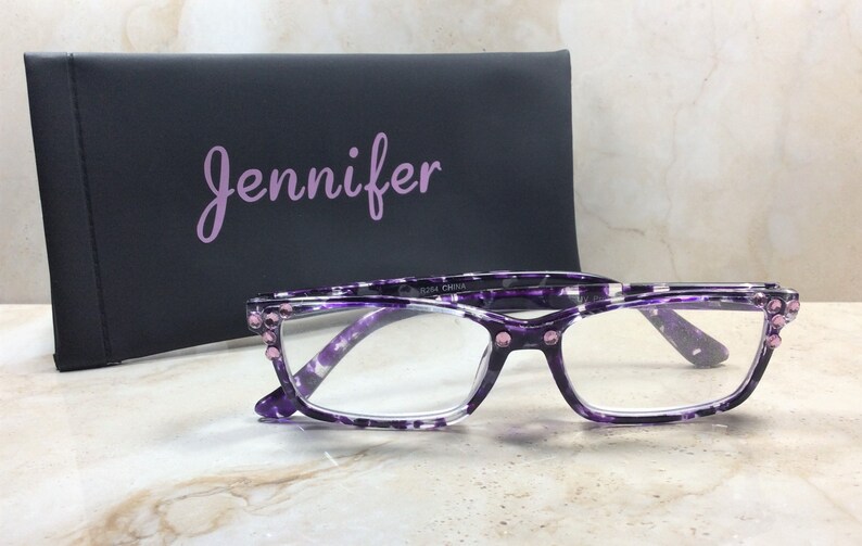 K&ouml;nnte beinhalten: Brille mit lila und wei&szlig;em Muster mit klaren Gl&auml;sern und rosa Edelsteinakzenten. Ein schwarzes Etui mit dem Namen "Jennifer" in lavendelfarbener Schrift befindet sich hinter der Brille. Die Brille liegt auf einer Marmoroberfl&auml;che.