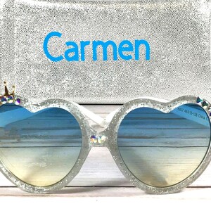 May include: Heart-shaped sunglasses with a glitter finish and a crown of rhinestones on one side. The sunglasses have a blue gradient lens and the name "Carmen" is embroidered on a silver pouch.