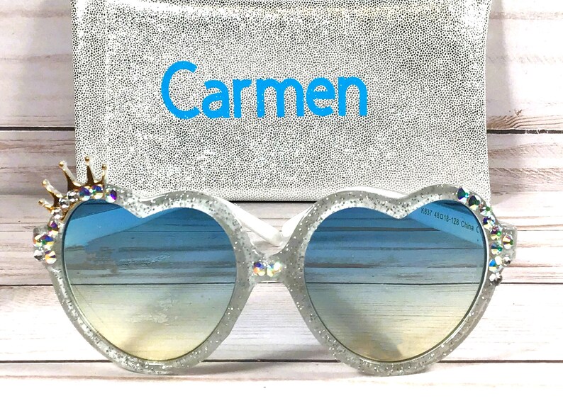 May include: Heart-shaped sunglasses with a glitter finish and a crown of rhinestones on one side. The sunglasses have a blue gradient lens and the name "Carmen" is embroidered on a silver pouch.