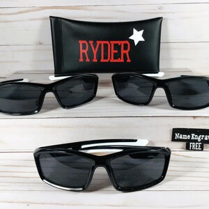 May include: Two pairs of black sunglasses with white accents and a black case with the word "RYDER" in red letters and a white star. The case also has the text "Name Engraved FREE".