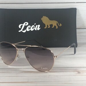May include: Aviator sunglasses with thin gold-tone metal frames and gradient lenses. The right lens features the white text Leon. Behind the sunglasses is a black rectangular case displaying the white text Leon and a gold lion silhouette icon.