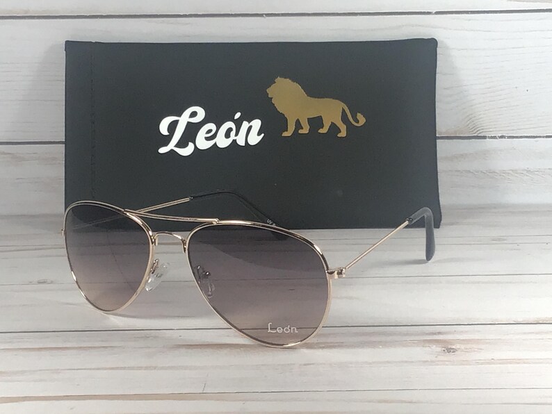 May include: Aviator sunglasses with thin gold-tone metal frames and gradient lenses. The right lens features the white text Leon. Behind the sunglasses is a black rectangular case displaying the white text Leon and a gold lion silhouette icon.