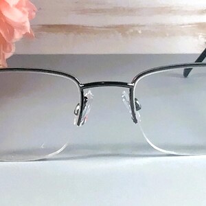 Pode incluir: &Oacute;culos sem aro com arma&ccedil;&atilde;o prateada e lentes transparentes. As hastes s&atilde;o pretas e adornadas com pequenos cristais brilhantes. Um arranjo floral rosa est&aacute; ao fundo, adicionando um toque de cor &agrave; imagem.