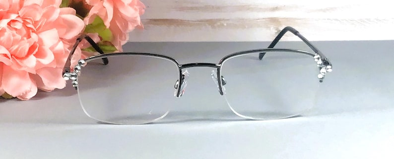 Pode incluir: &Oacute;culos sem aro com arma&ccedil;&atilde;o prateada e lentes transparentes. As hastes s&atilde;o pretas e adornadas com pequenos cristais brilhantes. Um arranjo floral rosa est&aacute; ao fundo, adicionando um toque de cor &agrave; imagem.