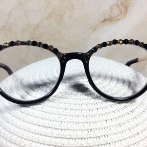 May include: Round, black-framed spectacles with clear lenses. The top of the frames are decorated with small, black and gold-coloured beads. The glasses are set against a white, textured background.
