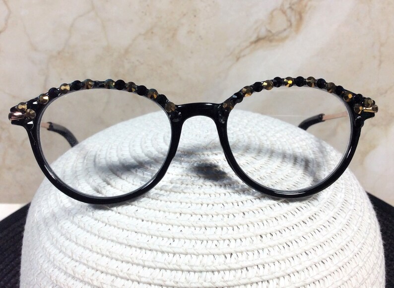 May include: Round, black-framed spectacles with clear lenses. The top of the frames are decorated with small, black and gold-coloured beads. The glasses are set against a white, textured background.