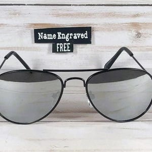 May include: Aviator sunglasses with thin black metal frames and reflective silver lenses. A small black rectangular sign positioned above the sunglasses features the white text Name Engraved FREE in a clear, sans-serif font style.