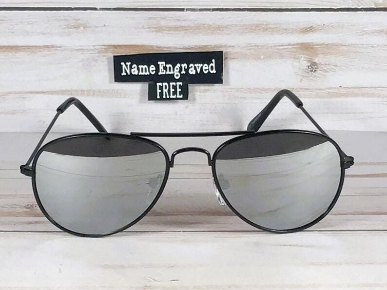 May include: Aviator sunglasses with thin black metal frames and reflective silver lenses. A small black rectangular sign positioned above the sunglasses features the white text Name Engraved FREE in a clear, sans-serif font style.