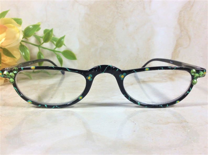 K&ouml;nnte beinhalten: Schwarz gerahmte Lesebrille mit floralem Design in T&uuml;rkis und Gelb. Die Fassungen sind mit gr&uuml;nen Strasssteinen an den Ecken verziert. Die Brille hat ein Halbrahmen-Design.