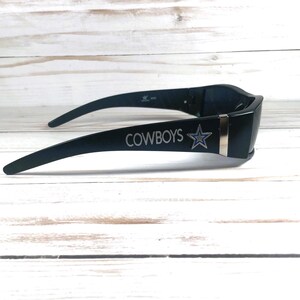 May include: Black sunglasses with the word "COWBOYS" and a star logo on the side. The sunglasses have a sleek, modern design with a dark frame and a silver accent.