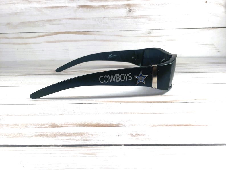 May include: Black sunglasses with the word "COWBOYS" and a star logo on the side. The sunglasses have a sleek, modern design with a dark frame and a silver accent.