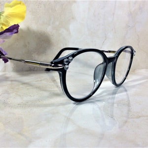 Puede incluir: Gafas con montura negra y lentes transparentes. Las monturas tienen pedrer&iacute;a decorativa en las esquinas y varillas plateadas. Las gafas est&aacute;n sobre un fondo claro, con una flor en la esquina superior izquierda.