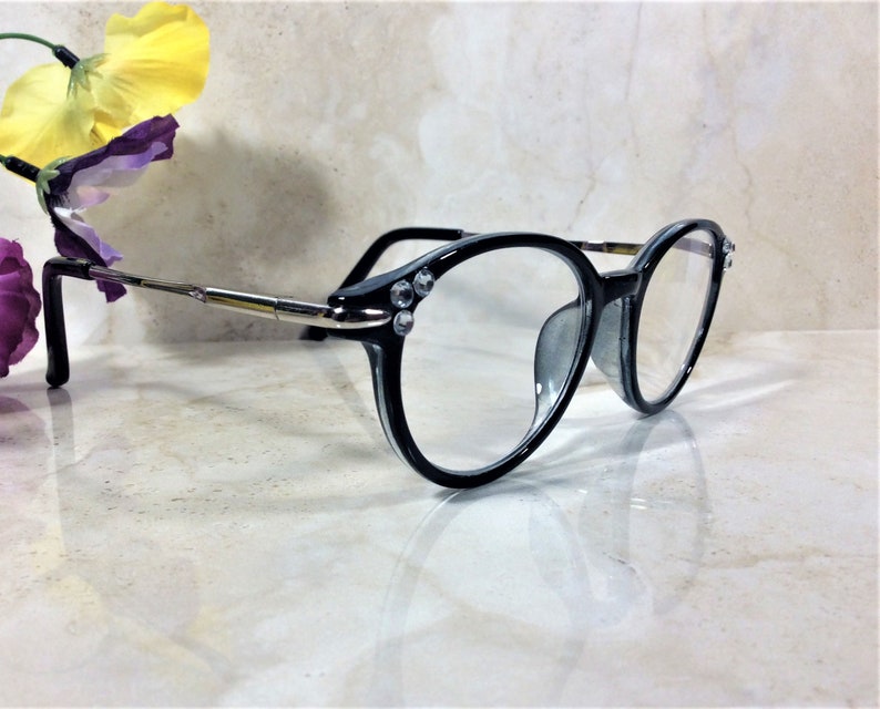 Puede incluir: Gafas con montura negra y lentes transparentes. Las monturas tienen pedrer&iacute;a decorativa en las esquinas y varillas plateadas. Las gafas est&aacute;n sobre un fondo claro, con una flor en la esquina superior izquierda.