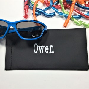 May include: A black personalized sunglasses case with the name "Owen" printed in white. The case is next to a pair of blue sunglasses with dark lenses.