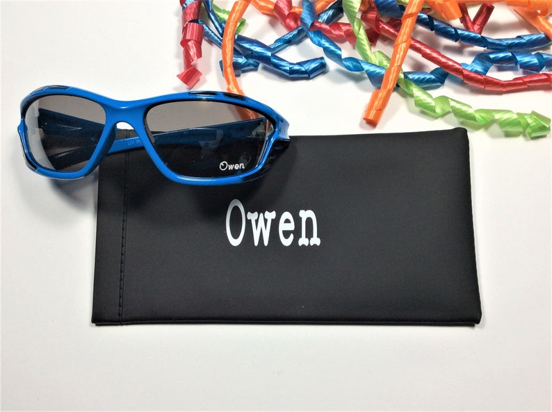 May include: A black personalized sunglasses case with the name "Owen" printed in white. The case is next to a pair of blue sunglasses with dark lenses.