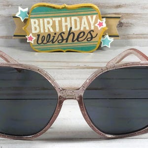May include: Pink glitter sunglasses with black lenses. The sunglasses have a square shape and a sparkly finish.