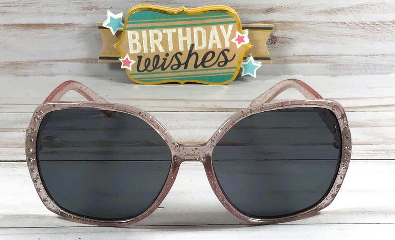 May include: Pink glitter sunglasses with black lenses. The sunglasses have a square shape and a sparkly finish.