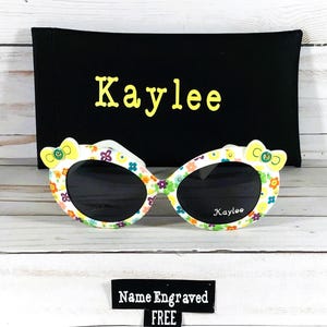 May include: White sunglasses with a floral pattern and yellow bows on the sides. The sunglasses come with a black carrying case with the name "Kaylee" embroidered on it. The sunglasses also have the name "Kaylee" printed on the lenses. A tag says "Name Engraved FREE".