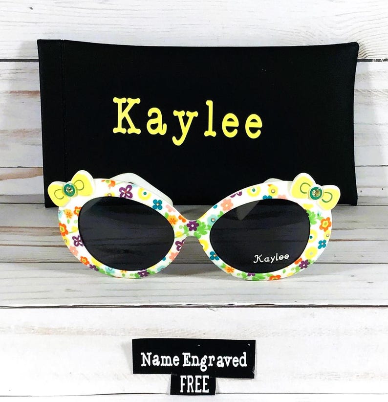 May include: White sunglasses with a floral pattern and yellow bows on the sides. The sunglasses come with a black carrying case with the name "Kaylee" embroidered on it. The sunglasses also have the name "Kaylee" printed on the lenses. A tag says "Name Engraved FREE".