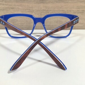 May include: Blue-framed spectacles with brown and white patterned arms. The frame has a rectangular shape with small, light blue embellishments on the temples. The glasses are set against a white surface.