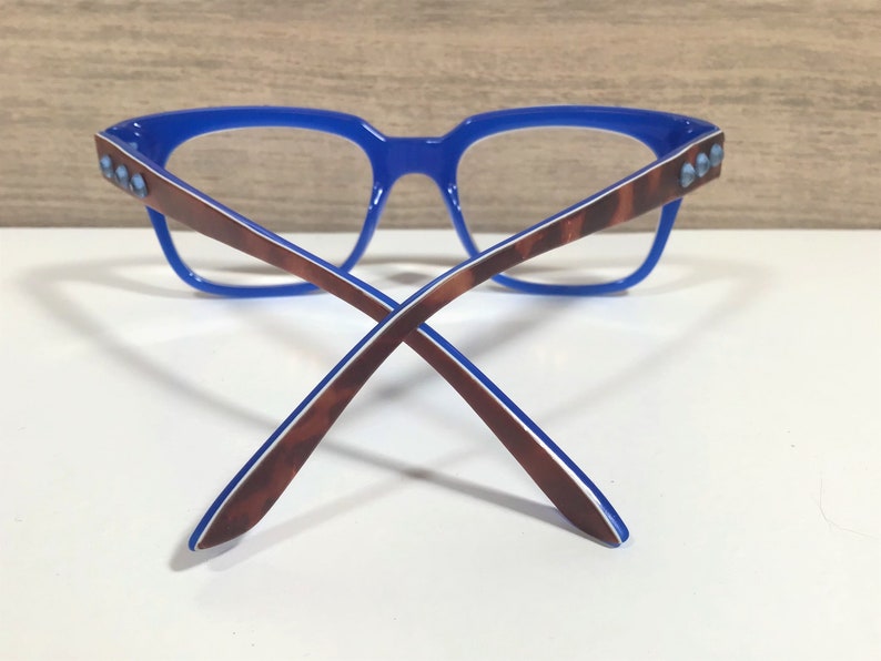 May include: Blue-framed spectacles with brown and white patterned arms. The frame has a rectangular shape with small, light blue embellishments on the temples. The glasses are set against a white surface.