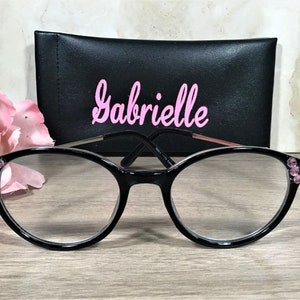 Puede incluir: Gafas negras con lentes transparentes y detalles de pedrer&iacute;a rosa en las monturas. Una funda negra con el nombre "Gabrielle" en escritura rosa est&aacute; detr&aacute;s de las gafas. Una flor rosa est&aacute; a la izquierda.