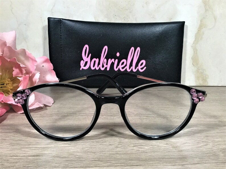 Puede incluir: Gafas negras con lentes transparentes y detalles de pedrer&iacute;a rosa en las monturas. Una funda negra con el nombre "Gabrielle" en escritura rosa est&aacute; detr&aacute;s de las gafas. Una flor rosa est&aacute; a la izquierda.